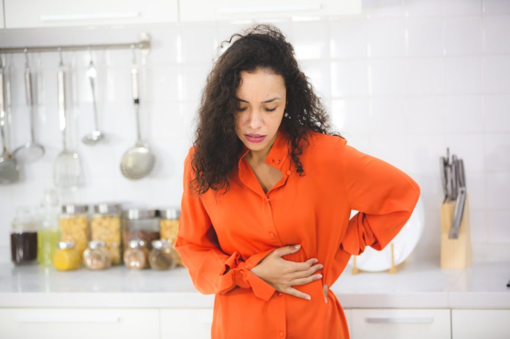 woman with curly hair standing in modern kitchen taking support of counter holding her abdomen hard