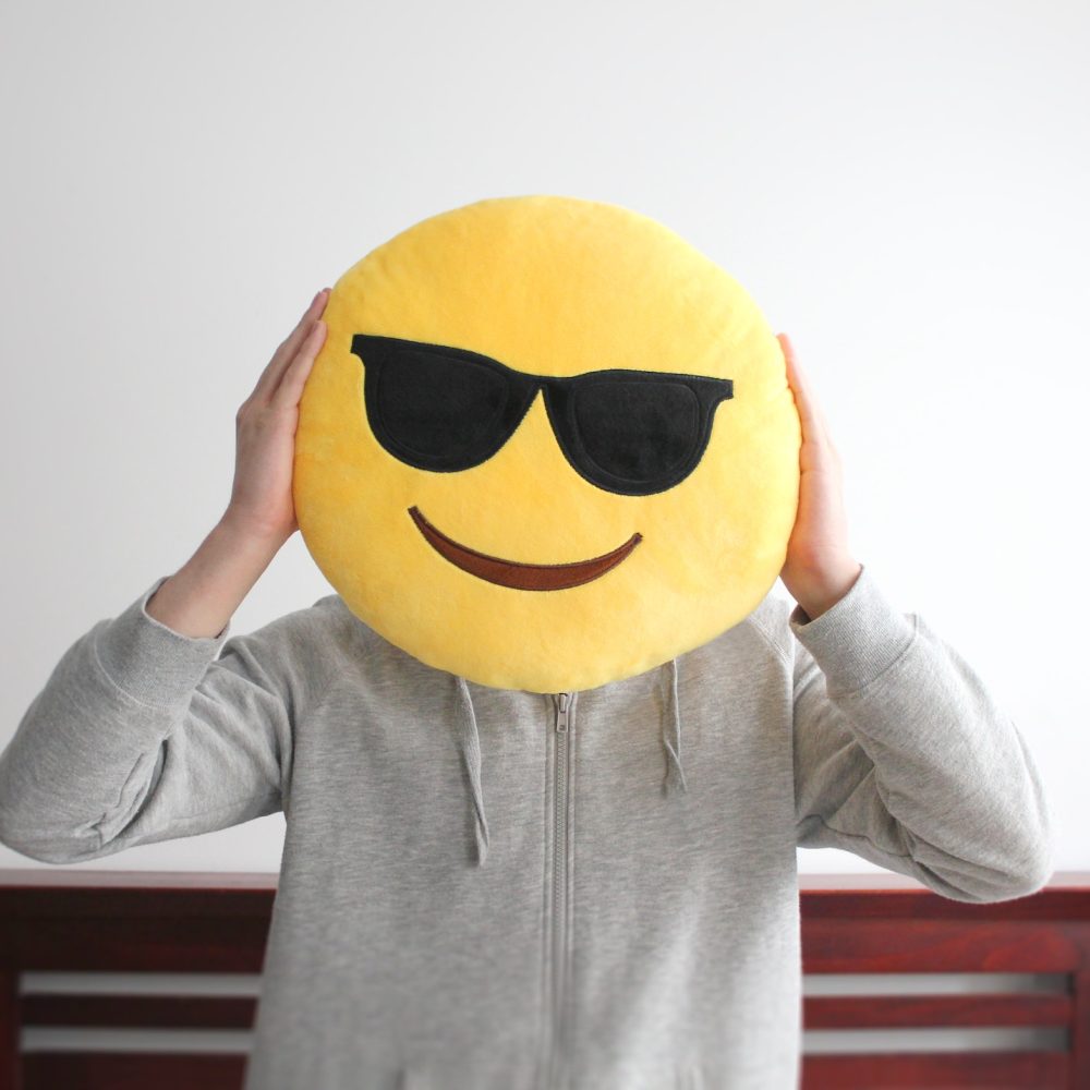 Bright yellow smiling emoji face wearing sunglasses cushion held up by a person